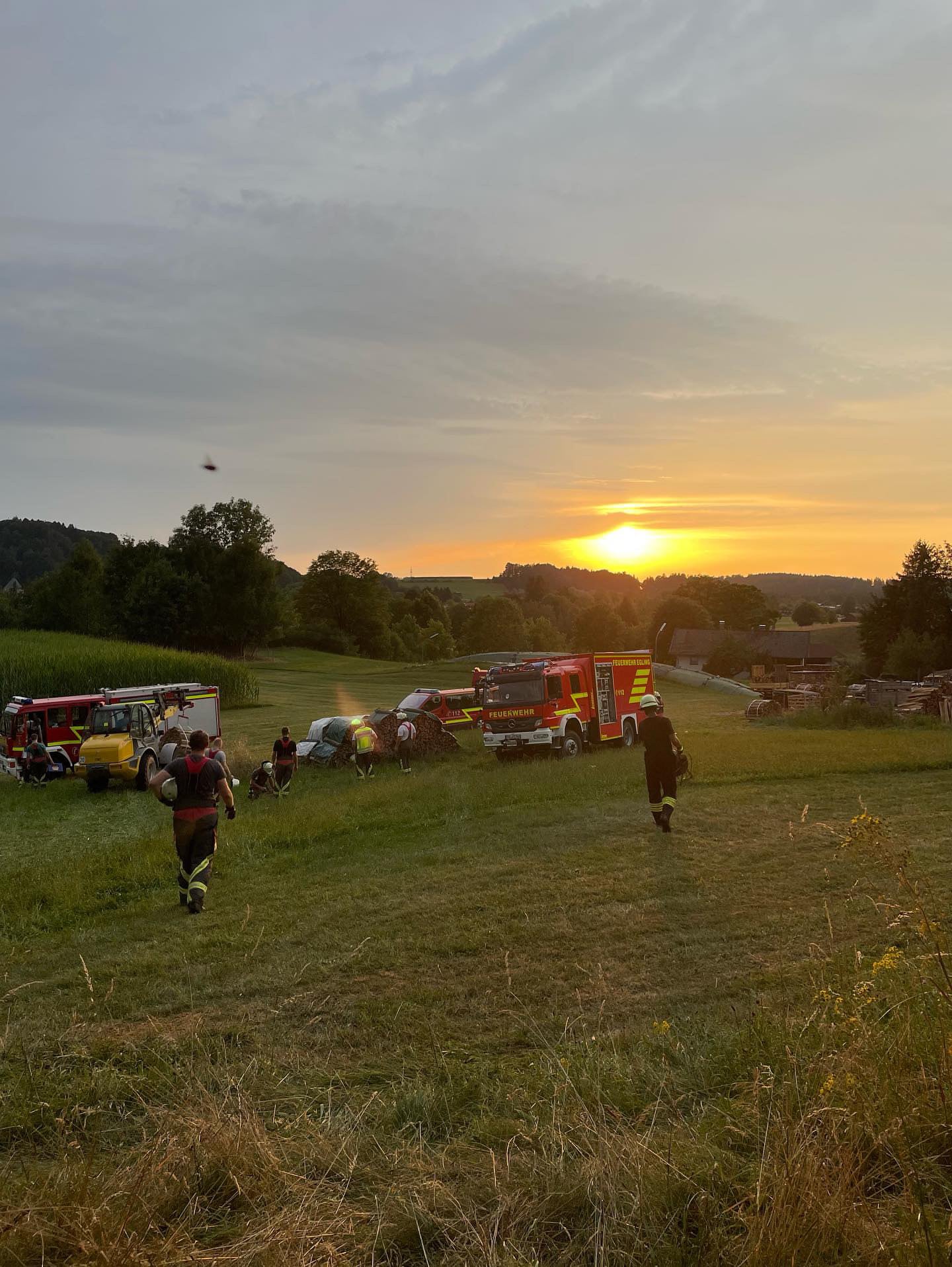 Sonnenuntergangsbild mit Feuerwehrfahrzeugen auf einer Ebene, mehrere Feuerwehrleute gehen von der Kamera weg, welliges Gelände und Wälder im Hintergrund
